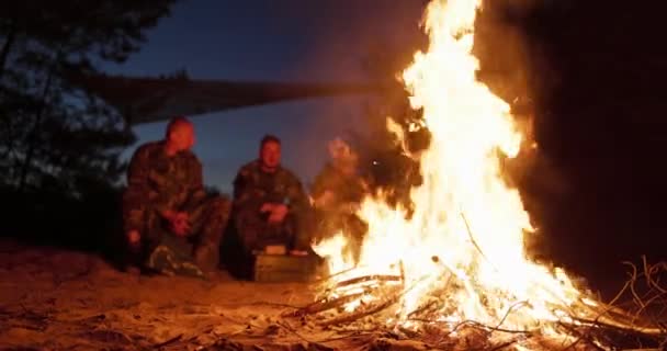Büyük bir şenlik ateşi, arkasında askeri üniformalı adamlar gece şöminenin başında oturuyor, konuşuyor ve kendilerini ısıtıyorlar. — Stok video