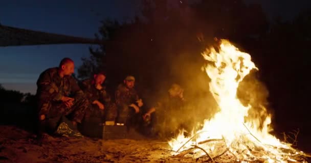 Uma grande chama de fogueira, atrás dela homens militares em uniformes de camuflagem estão sentados, conversando e aquecendo-se à noite pela lareira — Vídeo de Stock