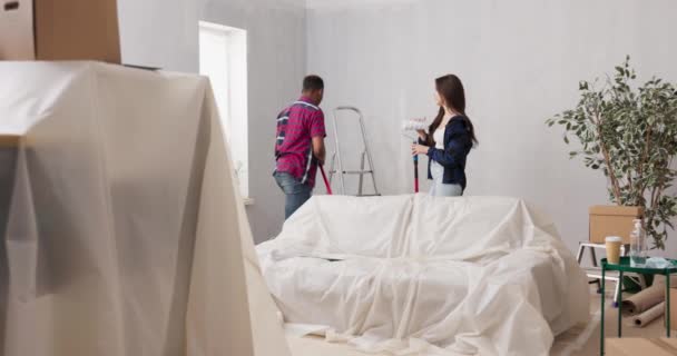 Védett bútoros lakás, védőfóliával borítva a festékfoltok ellen, felújítás, falak festése egy házaspár előtt, jegyesek, szerelmesek a festőhengerekbe — Stock videók
