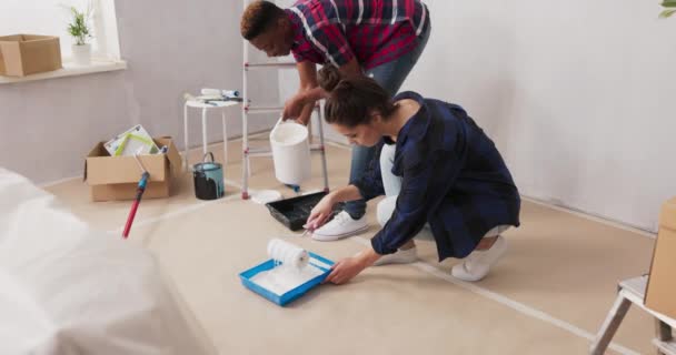 Två glada unga människor i kärlek renoverar sin första köpta lägenhet, de knäböjer på golvet förseglade med kartong scooping färg från färgbricka de håller rullar i händerna — Stockvideo