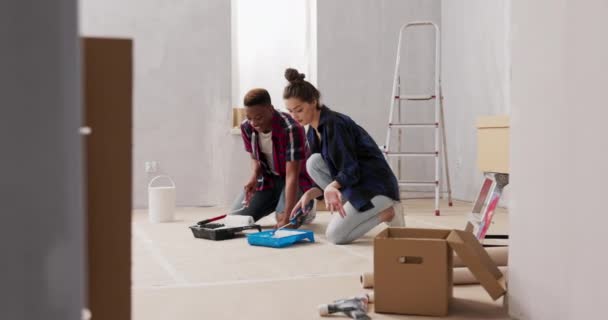 Dois jovens felizes no amor estão renovando seu primeiro apartamento comprado, eles estão ajoelhados no chão selado com papelão escavando tinta da bandeja de pintura que estão segurando rolos nas mãos — Vídeo de Stock