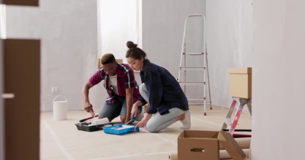 Přátelé spolu renovují byt, malují zdi v místnosti, muž ukazuje ženě, jak se dostat barvu na kolečko, aby se neušpinil, dívka se směje napodobuje chlapecké pohyby — Stock video