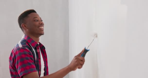 Young smiling laughing man dressed in jeans and checkered shirt works as wall painter during renovations of apartments houses he holds roller on handle — Stock Video
