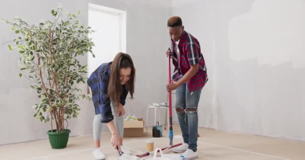 Dois jovens pessoas alegres trabalhando juntos para terminar um apartamento segurar rolos de tinta em suas mãos ficar sujo com tinta branca em torno deles caixas escada amigos pintura paredes — Vídeo de Stock