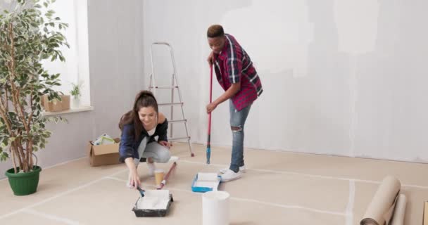 Twee jonge vrolijke mensen die samenwerken om een appartement af te maken houden verfrollen in hun handen vuil met witte verf rond die dozen ladders vrienden schilderen muren — Stockvideo