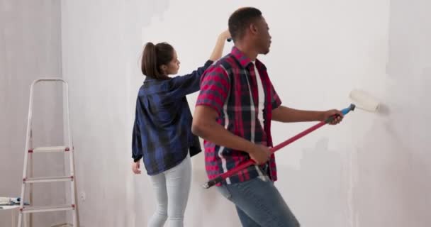 Glimlachende vrolijke vrienden die plezier hebben terwijl ze een appartement renoveren knappe man die rond loopt met een vrouw met een roller bedekt met verf ze schilderen spinnen hun heupen zingend dansen — Stockvideo