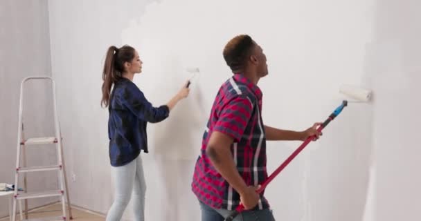 Sorrindo amigos alegres se divertindo ao renovar terminar um apartamento bonito homem brincando com a mulher segurando rolo coberto de tinta que pintam girar seus quadris cantando dança — Vídeo de Stock