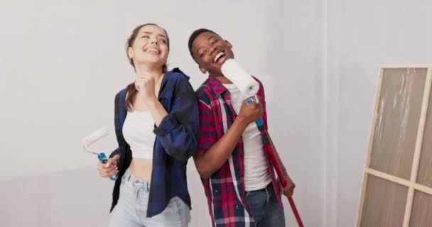 Entreter amigos alegres passar o tempo juntos, se divertir ao renovar o quarto, apartamento, segurar rolos de tinta em suas mãos, cantando — Vídeo de Stock