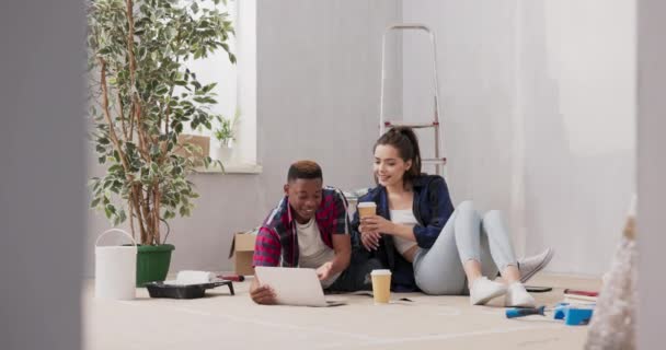 Pareja está pasando tiempo en el nuevo apartamento redecorar la sala de pintura paredes pintura accesorios dispersos alrededor de los amantes está navegando a través de inspiraciones en el ordenador portátil ordenar muebles, beber café para ir — Vídeos de Stock