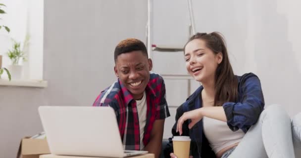 Casal está gastando tempo em novo apartamento redecorando paredes de pintura quarto pintura acessórios espalhados ao redor amantes está navegando através de inspirações no laptop rindo beber café para ir — Vídeo de Stock