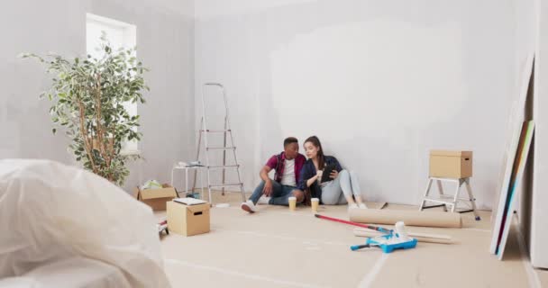 Bonito homem senta-se no chão com sua bela namorada, fazendo uma pausa de paredes de apartamentos de pintura durante uma renovação, bebendo café take-out, navegando inspiração em um tablet — Vídeo de Stock