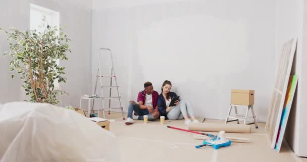 Όμορφος άνθρωπος κάθεται στο πάτωμα με την όμορφη αγαπημένη του, κάνοντας ένα διάλειμμα από τη ζωγραφική τοίχους διαμέρισμα κατά τη διάρκεια μιας ανακαίνισης, πίνοντας take-out καφέ, περιήγηση έμπνευση σε ένα tablet — Αρχείο Βίντεο