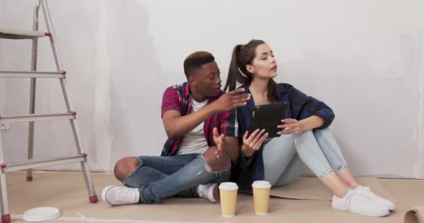 Pareja enamorada sentarse en el suelo durante el descanso de la pintura de las paredes del apartamento durante la renovación beber café para llevar navegación inspiración en la tableta averiguar detalles de paisajismo sonrisa para la cámara — Vídeo de stock