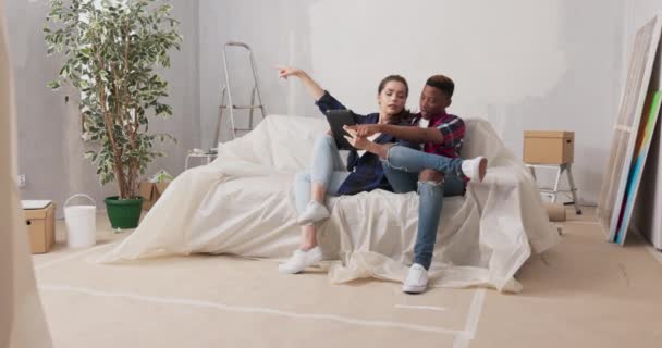 Különböző nemzetiségű párok ülnek a kanapén a nappaliban, az újonnan vásárolt lakásban felújítás alatt, szerelmesek megvitatják dekoráció bútor falfesték nézés projektek tabletta — Stock videók