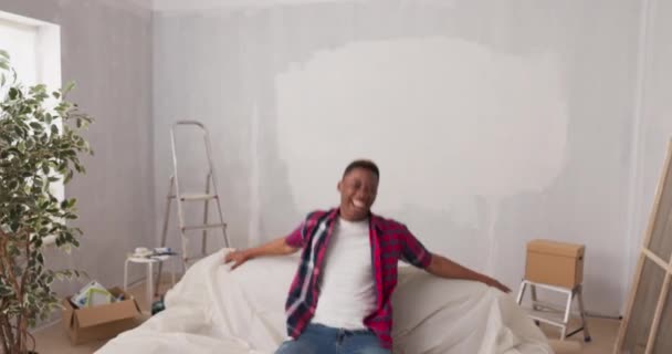 En man tittar på kameran med ett stolt ansikte och ler. I bakgrunden finns en vägg målad i en nyrenoverad lägenhet glad trött pojke sitter på säkrad soffa — Stockvideo