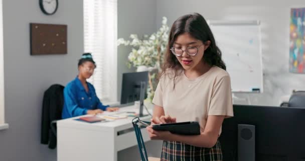 La responsable marketing, une belle fille avec des lunettes, tient une tablette dans sa main, vérifie la portée sur les médias sociaux, discute avec un collègue de l'entreprise des résultats, des progrès — Video