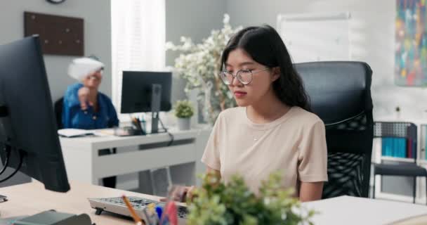 Οι υπάλληλοι του γραφείου κάθονται σε γραφεία μπροστά από υπολογιστές κοροϊδεύοντας μια γυναίκα με κορεάτικη ομορφιά εργάζεται επιμελώς ένα άλλο κορίτσι ενοχλεί — Αρχείο Βίντεο
