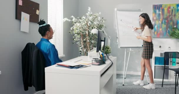 Dos mujeres en la oficina corporativa en el descanso una de las mujeres está de pie en la pizarra con el marcador en la mano, están de acuerdo en hacer bromas y tachar las líneas, cambiar la apariencia del dibujo se ríen chocan — Vídeo de stock