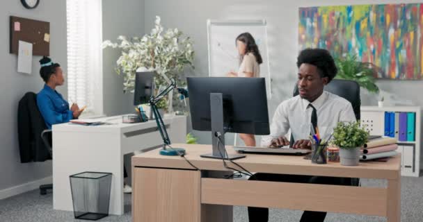 Egy fehér inges és afro fejű férfi koncentrációban dolgozik egy számítógépen a háttérben, női munkatársak dolgoznak egy projekten, statisztikákat készítenek egy grafikon rajzolásával, jegyzetelnek. — Stock videók