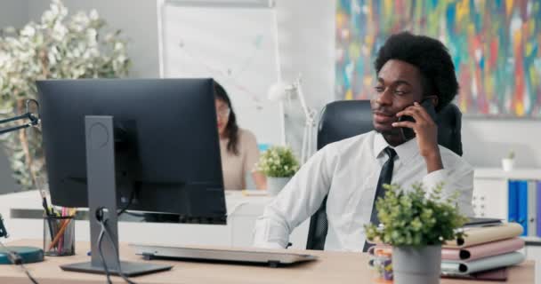 Enfocado, hombre serio con un afro en la cabeza, con una camisa y corbata, se sienta en la oficina hablando por teléfono de negocios, el cuidado de los negocios de la empresa, la organización de un evento, conferencia, formación para los empleados — Vídeos de Stock