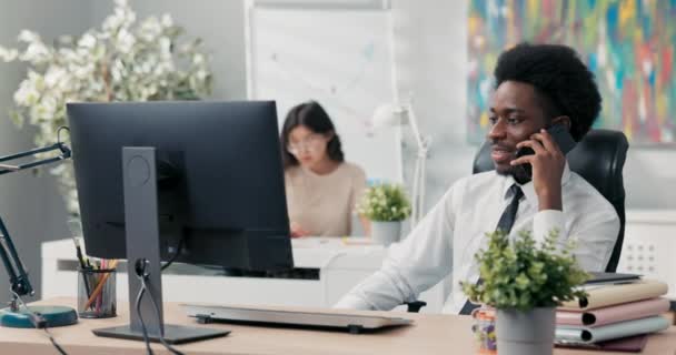 Skupiony, poważny mężczyzna z afro na głowie, ubrany w koszulę i krawat, siedzi w biurze i rozmawia przez telefon służbowy, dba o interesy firmy, organizuje imprezę, konferencję, szkolenia dla pracowników — Wideo stockowe