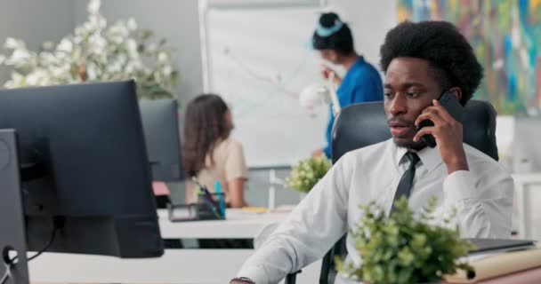 穿着白衬衫、打着黑色领带、头戴非洲裔经理的男人面带微笑坐在办公桌前，手里拿着电脑，手持电话号码打了个电话 — 图库视频影像