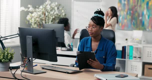 Atrakcyjna dziewczyna w średnim wieku siedzi na krześle w biurze firmy przy biurku przed komputerem, trzymając tablet w rękach, dbając o obowiązki, odpowiadając na maile, wysyłając wiadomości, sprawdzając oferty — Wideo stockowe
