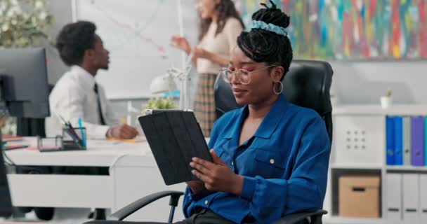 En kvinna i blå skjorta och glasögon sitter i en bekväm stol på jobbet, håller en tablett i händerna, bläddrar i sociala nätverk, svarar på meddelanden — Stockvideo