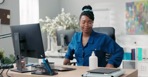 Una mujer atractiva con camisa y gafas viene a trabajar, se sienta en el escritorio, desinfecta sus manos con un líquido antes de hacer clic en los dedos en el teclado de la computadora, la salud y la seguridad de la empresa — Vídeos de Stock