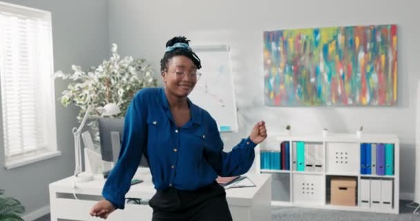 Mavi gömlekli ve şık gözlüklü zarif bir kız, ofisin ortasında dans ediyor, kollarını öne doğru sallıyor, şirketin başarısı için mutlu, yüzünde gülümseme olan bir iş kadını. — Stok video