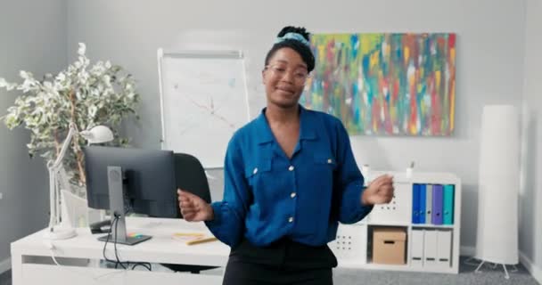 Mavi gömlekli ve şık gözlüklü zarif bir kız, ofisin ortasında dans ediyor, kollarını öne doğru sallıyor, şirketin başarısı için mutlu, yüzünde gülümseme olan bir iş kadını. — Stok video