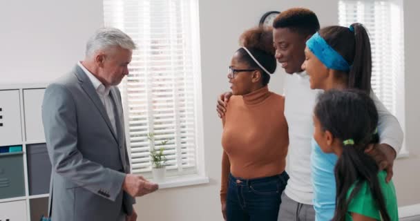 Starszy mężczyzna z siwymi włosami, agent nieruchomości sprzedaje mieszkanie młodej parze z dziećmi, wręcza kobiecie klucze wyjaśniające szczegóły, rodzina przytula się ze szczęściem — Wideo stockowe