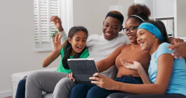 A happy family sits on the couch, relaxing, spending time together in the afternoon, parents with two daughters waving at the tablet camera, having a video conversation with friends — Stock Video
