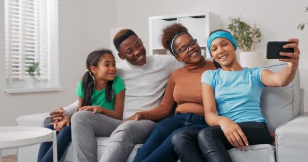 Una niña en una blusa azul y diadema se sienta en el sofá con su familia, se toman una selfie con un teléfono inteligente, sonríen para la cámara, eligen la mejor foto y la publican en las redes sociales — Vídeos de Stock