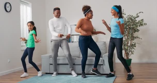 Tiempo en familia pasado en casa, alegre, pareja loca junto con sus hijas bailan en el centro de la sala de estar, juegan, fiesta, cantar, saltar, divertirse juntos — Vídeo de stock