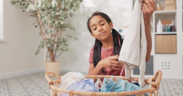 En leende söt flicka sitter i mitten av badrummet, tvättstuga, framför en stor korg fylld med färgglada kläder i händerna, dotter hjälpa mamma med hushållssysslor — Stockvideo