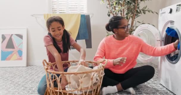 뜨개 질을 하는 여자와 딸 이 빨래 바구니에서 옷을 꺼내 세탁기의 드럼통에 싣고 집안일을 돕고 있다 — 비디오