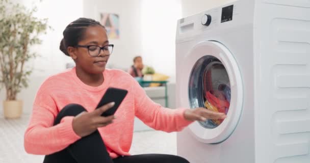 Multifunctional washing machine controlled by mobile app, fast program, woman closes drum with colorful clothes, taps fingers on phone, walks away and sits on couch next to daughter — Stock Video