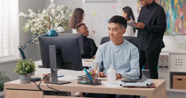 Un hombre de belleza coreana asiática se sienta frente a una computadora de oficina, usando un navegador web, trabajando frente a una pantalla, manejando las redes sociales de las empresas, creando anuncios, publicaciones, gráficos — Vídeos de Stock