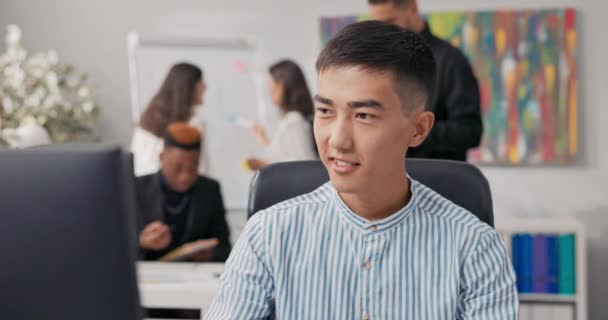 Um homem de beleza asiática coreana senta-se na frente de um computador de escritório, usando um navegador da web, trabalhando na frente de uma tela, manipulando as mídias sociais da empresa, criando anúncios, posts, gráficos — Vídeo de Stock