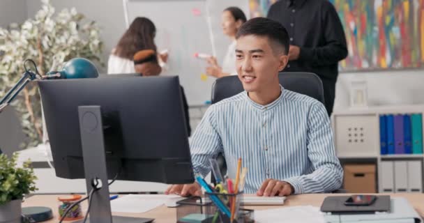 Mężczyzna azjatycko-koreańskiej urody siedzi na komputerze biurowym, za pomocą przeglądarki internetowej, pracując przed monitorem, uczy się dobrych informacji przepełniony radości odwraca się i pługa przez to, szczęście — Wideo stockowe