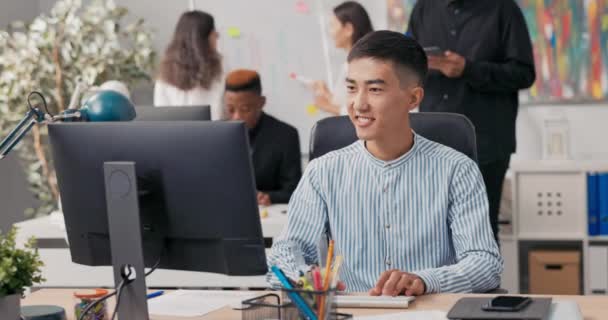 Ein Mann asiatisch-koreanischer Schönheit sitzt auf einem Bürocomputer, bedient einen Webbrowser, arbeitet vor einem Monitor, lernt gute Informationen, dreht sich überglücklich um und pflügt durch ihn, Glück — Stockvideo