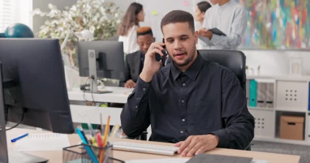 Hombre vestido con una elegante camisa negra se sienta en el escritorio frente a un ordenador, recibe una llamada en el teléfono inteligente, explica el problema con la calma, habla con el que llama, proporciona información importante — Vídeos de Stock