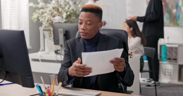 O funcionário de escritório senta-se na frente do computador na mesa tem chamada de vídeo, conexão com a internet através da webcam, mostra documento em papel com faturamento que o cara explica ao colega de trabalho — Vídeo de Stock