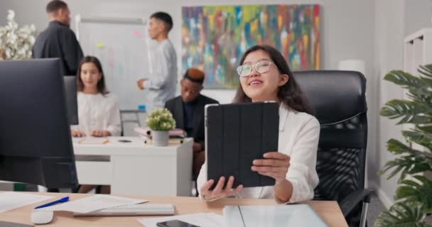 Mujer en camisa blanca y gafas se sienta en el escritorio sosteniendo la tableta en las manos teniendo video conversación con el jefe a través de webcam compañeros de trabajo de pie en la pizarra en el fondo y saludar a la ola — Vídeos de Stock