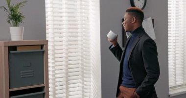 Ciddi, odaklanmış, düşünceli bir şirket başkanı, bir işadamı pencerenin önünde dikiliyor, panjurlarla kaplı ve içine bakıyor, iş hakkında düşünüyor, kahve içiyor.