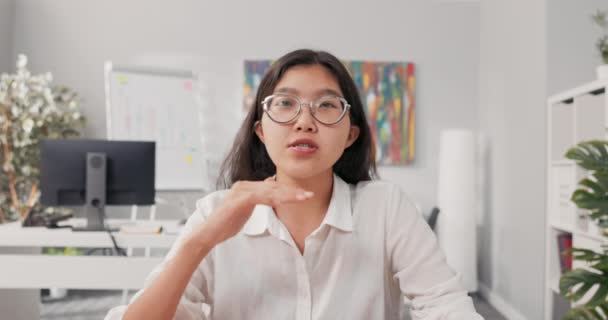 Uma mulher asiática coreana sorridente bonita com óculos senta-se em um escritório vestido com uma camisa branca esperta, uma mulher de negócios prende uma chamada vídeo, conferências, faz suas perguntas do chefe sobre o Internet — Vídeo de Stock