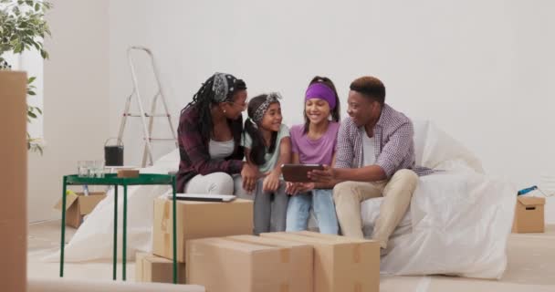 Mutlu bir aile, yenileme sırasında evdeki kanepede oturur ve iki kızı boya renkleri için odaları nasıl bitireceklerine dair ilham tabletine göz atarlar. — Stok video