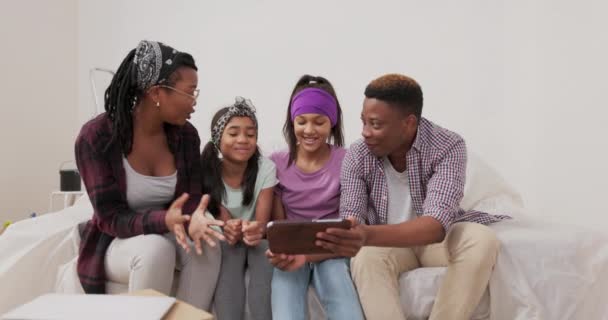 Μια ευτυχισμένη οικογένεια κάθεται στον καναπέ στο διαμέρισμα κατά τη διάρκεια της ανακαίνισης παντρεμένο ζευγάρι και οι δύο κόρες τους περιηγηθείτε στο tablet για έμπνευση για το πώς να τελειώσει τα δωμάτια που αναζητούν χρώματα χρώμα — Αρχείο Βίντεο