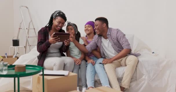 Uśmiechnięta para z dwiema córkami siedzi na kanapie w odnowionym mieszkaniu kobieta trzyma tablet w rękach robi sobie selfie ze swoimi bliskimi śmiejąc się żartując nieudana fotografia — Wideo stockowe